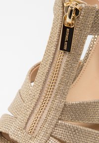 Gros plan d’une sandale beige tissée avec une fermeture éclair dorée et une languette gravée du nom de la marque Michael Kors.