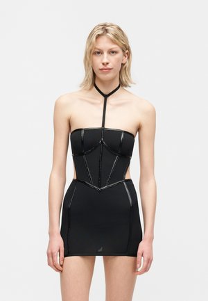 Szőke nő fekete, váll nélküli miniruhában kivágásokkal, egy függőleges pánttal összekötött gallérral és kontrasztos varrási részletekkel.