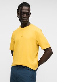 Κίτρινο βαμβακερό t-shirt με κοντά μανίκια, λαιμόκοψη τύπου crew και μικρό κεντητό λογότυπο στο στήθος. Συνδυάζεται με μπλε παντελόνι.