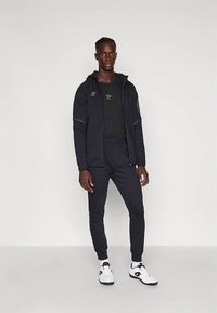 Homem alto de pé, vestindo roupas desportivas pretas, um hoodie com fecho, calças de treino e sapatilhas brancas com solas pretas.