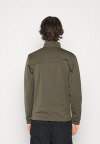 Olivově zelená fleecová bunda s vysokým límcem, dlouhými rukávy a černým logem na horní části zad. Hladká, texturovaná tkanina.