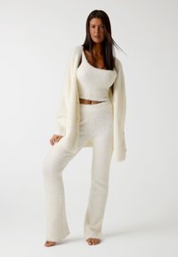 Completo in due pezzi in tessuto bianco soffice; presenta un top corto e pantaloni a zampa abbinati a un cardigan lungo. Bordo arrotondato, texture morbida.