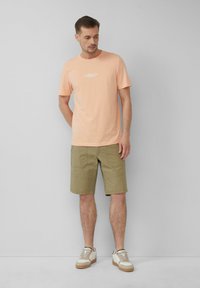 T-shirt di cotone pesco con logo bianco, abbinata a pantaloni cargo verde oliva con tasche a toppa e sneaker beige con suole testurizzate.