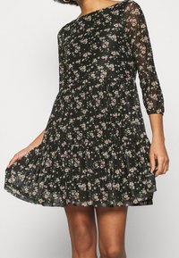 Robe noire à fleurs avec des manches trois-quarts en dentelle transparente, un bas plissé et de petits motifs floraux roses et verts tout au long. Tissu doux et léger.