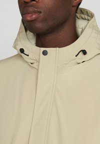 Chaqueta ligera beige con cuello alto, presenta herrajes negros y cordones ajustables en el cuello. Textura suave con costuras sutiles.