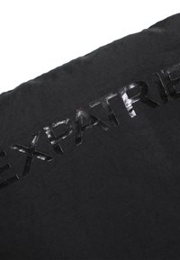 Sort stofpude med glinsende sort skrift, der staver "EXPATRIÉ" i midten. Overfladen har en glat tekstur.