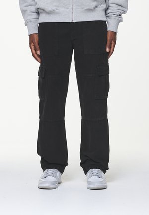 Zwarte cargobroek van stof, met meerdere zakken, een ontspannen pasvorm en een iets losse uitstraling. Gepaard met grijze sneakers.