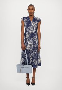Rochie bleu închis cu un model de palmieri albi, cu mâneci scurte, talie prinsă cu un brâu și fustă fluidă. Este însoțită de o geantă bleu deschis și tocuri negre.