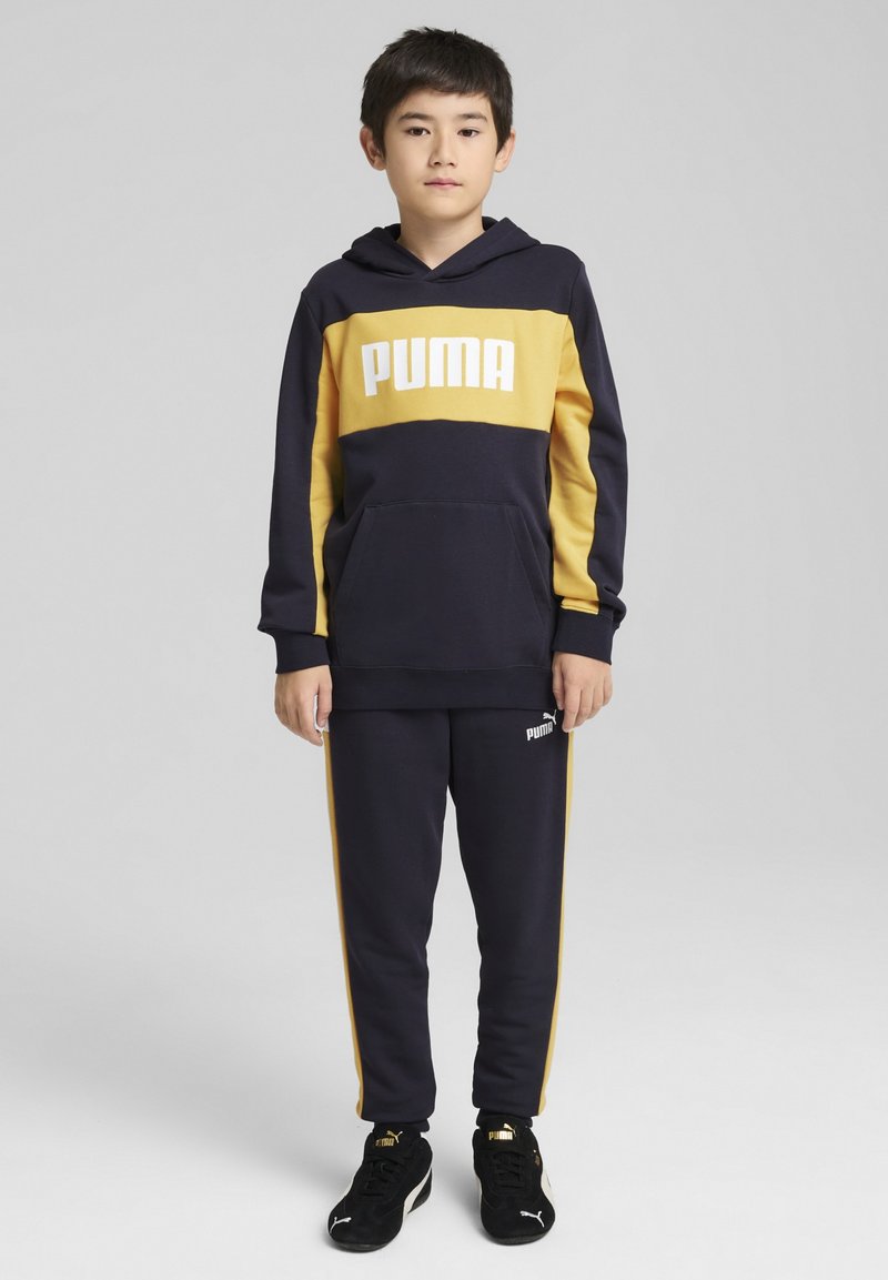Jūrų mėlynos ir geltonos PUMA treningo komplektas su gobtuvu, kuriame yra megztinis ir kelnes. Pasižymi baltu logotipu, šoninėmis juostelėmis ir minkšta medžiaga.