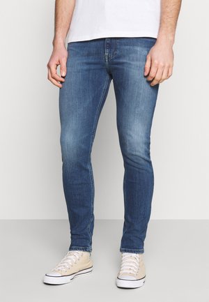 Vaqueros slim fit de color azul oscuro con un lavado deslavado, que cuentan con cinco bolsillos y trabillas para el cinturón, combinados con zapatillas de lona de color claro.