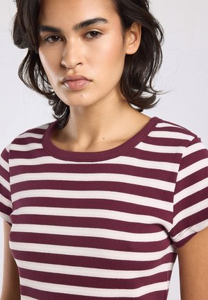 Jonge vrouw met kort donker haar die een bordeauxrode en witte gestreepte top met korte mouwen draagt, kijkt met een neutrale uitdrukking naar voren.