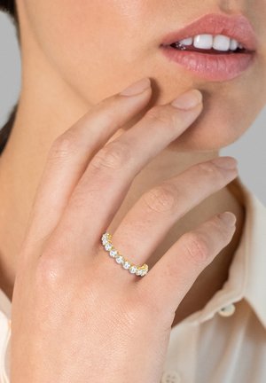 Anello in oro con pietre chiare e gialle alternate, caratterizzato da una texture liscia e un design curvato, indossato su una mano vicino al viso.
