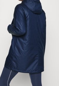 Marineblauwe, gevoerde jas met een gladde textuur, met een lange, rechte snit. Elastische manchetten en zijzakken zijn opgenomen in het ontwerp.