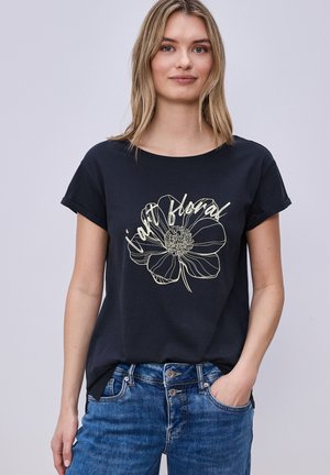 Ung kvinde iført en sort t-shirt med en stor konturtegnede blomst og ordet "plant floral" kombineret med blå jeans.