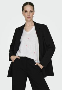 Blazer noir sur une chemise blanche à col en V avec des impressions de cœurs rouges, associé à un pantalon noir ; tissu lisse et coupe ajustée.
