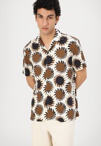 Camicia a maniche corte con sfondo crema e grandi motivi di sole marroni e neri, chiusura a bottoni e colletto appuntito.