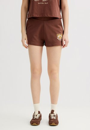 Mujer con pantalones cortos marrones con un gráfico de sol en la pierna izquierda, camiseta marrón a juego, calcetines blancos y zapatillas marrones, de pie sobre fondo blanco.