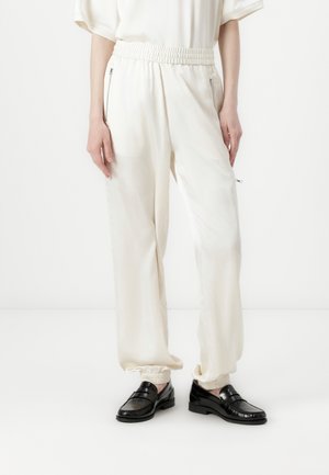 Cuffed, cream-colored trousers with a shiny finish, featuring an elastic waistband and side zip pockets. Paired with black loafers.