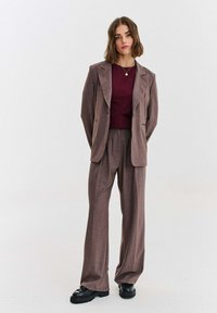 Brązowy garnitur w kratę z dopasowaną marynarką i spodniami o szerokich nogawkach, zestawiony z bordową bluzką. Całość uzupełniają czarne, masywne buty.