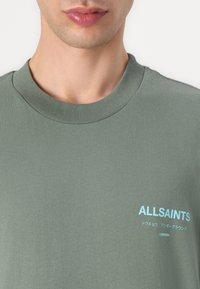 Zaļš kokvilnas T-krekls ar ribotu apkakli un gaiši zilu logo druku, kas lasāma "ALLSAINTS LONDON" un papildu tekstu japāņu valodā.