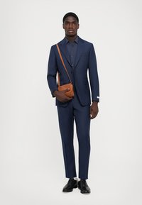 Marineblå skræddersyet jakkesæt med en tekstureret finish, kombineret med en mørk skjorte og en brun læder crossbody taske. Sorte dressko fuldender looket.