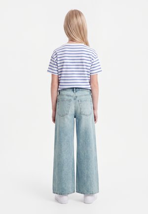 Barn med langt blond hår står med ryggen til iført en blå og hvidstribet t-shirt, vide lyse denimjeans og hvide sko.