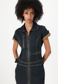 Denim jumpsuit med korte ermer, mørkeblå farge, glidelås foran og to brystlommer. Har teksturert stoff og spenne accenter i midjen.