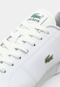 Sapatilhas brancas em couro com atacadores brancos, apresentam um logo de crocodilo verde da Lacoste e detalhes de costura subtil nas laterais.