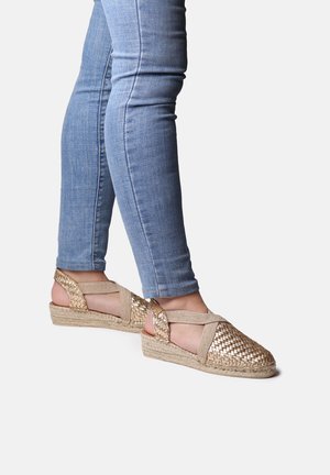 Woven gold espadrille with crisscross straps, jute sole, and open heel. Paired with blue skinny jeans, showcasing a casual style.