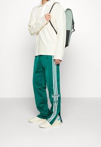 Osoba v tyrkysových teplákových kalhotách Adidas s bílými pruhy a patentkami, bílých teniskách, smetanovém tričku s dlouhým rukávem a nese zelený batoh.