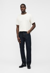 Mężczyzna stojący, ubrany w gładki biały podkoszulek z krótkim rękawem, ciemnoniebieskie dżinsy i czarne eleganckie buty, z jedną ręką w kieszeni.