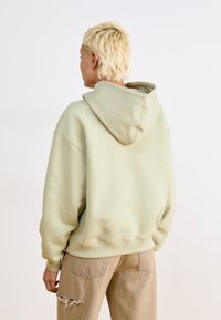 Sweatshirt oversized de cor verde claro com capuz ajustável, punhos e bainha canelados. Usado com calças bege com acabamentos desgastados.