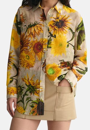 Langærmet skjorte med knapper og store gule solsikkeprint, båret med en beige mininederdel, en hånd i lommen.