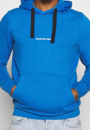 Osoba w jaskrawo niebieskiej bluzie z kapturem z czarnymi troczkami i napisem "REDEFINED REBEL", ręce w przedniej kieszeni, widoczne jasne, sprane dżinsy.