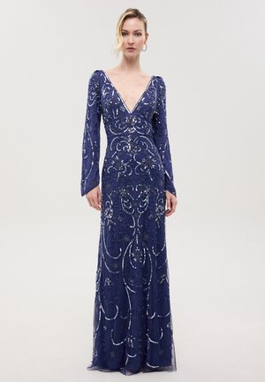 Vrouw draagt een marineblauwe jurk met lange mouwen met zilveren kraal- en paillettenbloemenpatronen en een diepe V-hals, staande tegen een eenvoudige lichte achtergrond.
