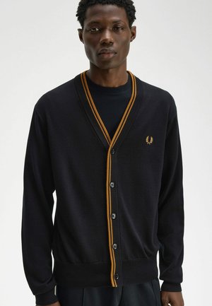 Uomo che indossa un cardigan nero con bordi e bottoni arancioni sopra una camicia nera, in piedi davanti a uno sfondo chiaro e uniforme.