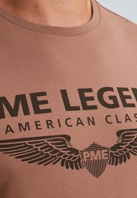 Hnědé bavlněné tričko s velkým černým grafickým motivem s křídly a textem "PME LEGEND AMERICAN CLASSIC." Design s kulatým výstřihem, hladká textura.