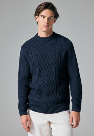 Granatowy sweter z dzianiny z wzorem w warkocz, teksturowanym korpusem oraz ściągaczami przy mankietach i hemie. Model ma na sobie kremowe spodnie, stoi na neutralnym tle.