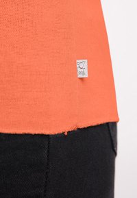 Korte mouw oranje gebreide top met een rauwe zoom en een geweven label met een klein hondenafbeelding, in combinatie met donkere denimstof.