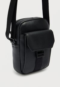 Μαύρο δερμάτινο τσαντάκι χιαστί με υφή στην επιφάνεια, κλείσιμο με καπάκι, ρυθμιζόμενο λουρί και διακριτική λεπτομέρεια με λογότυπο στο μπροστινό μέρος.