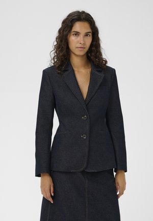 Lokkis juustega naine, kes kannab tumedast denimist kahe nupuga bleiserit ja sobivat tumedat denimi seelikut, seisab lihtsa taustaga ees.