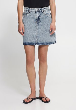 Frau trägt einen hellblauen Acid-Wash Denim-Minirock, schwarzen Oberteil, silberne Ringe und schwarze Flip-Flops, steht vor einem einfarbigen Hintergrund.