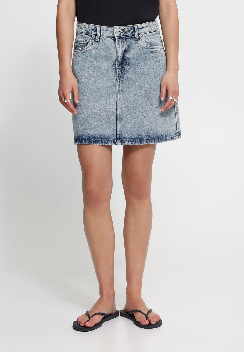 Femme portant une mini-jupe en jean bleu clair acidulé, un top noir, des bagues argentées et des tongs noires, debout devant un fond uni.