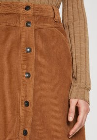 Bruine ribfluwelen rok met verticale rib structuur en zwarte knoopsluiting. De stof heeft een zachte uitstraling met zichtbare stiksels.