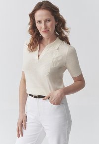 Polo shirt beige lavorato a maglia con maniche corte, caratterizzato da un design floreale ricamato in bianco sul petto, abbinato a jeans bianchi e una cintura marrone.