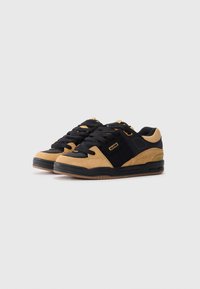 Une paire de baskets basses noires et beiges avec des lacets noirs, des semelles épaisses et des étiquettes de marque sur un fond blanc uni.