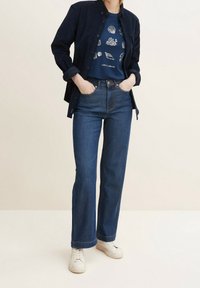 Marinblå kordflads skjorta över en blå grafisk t-shirt med vita snäcka-mönster, kombinerat med mörk denim med utsvängda byxor och vita sneakers.