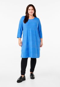 Abito a maglia bleu a righe con maniche a tre quarti, caratterizzato da diverse trame testurizzate. Abbinato a pantaloni neri aderenti e scarpe nere spesse.