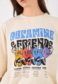 T-shirt de algodão bege com personagens de cartoon coloridos (vermelho, amarelo, azul, roxo) com expressões faciais distintas e positivo, e texto em negrito abaixo.