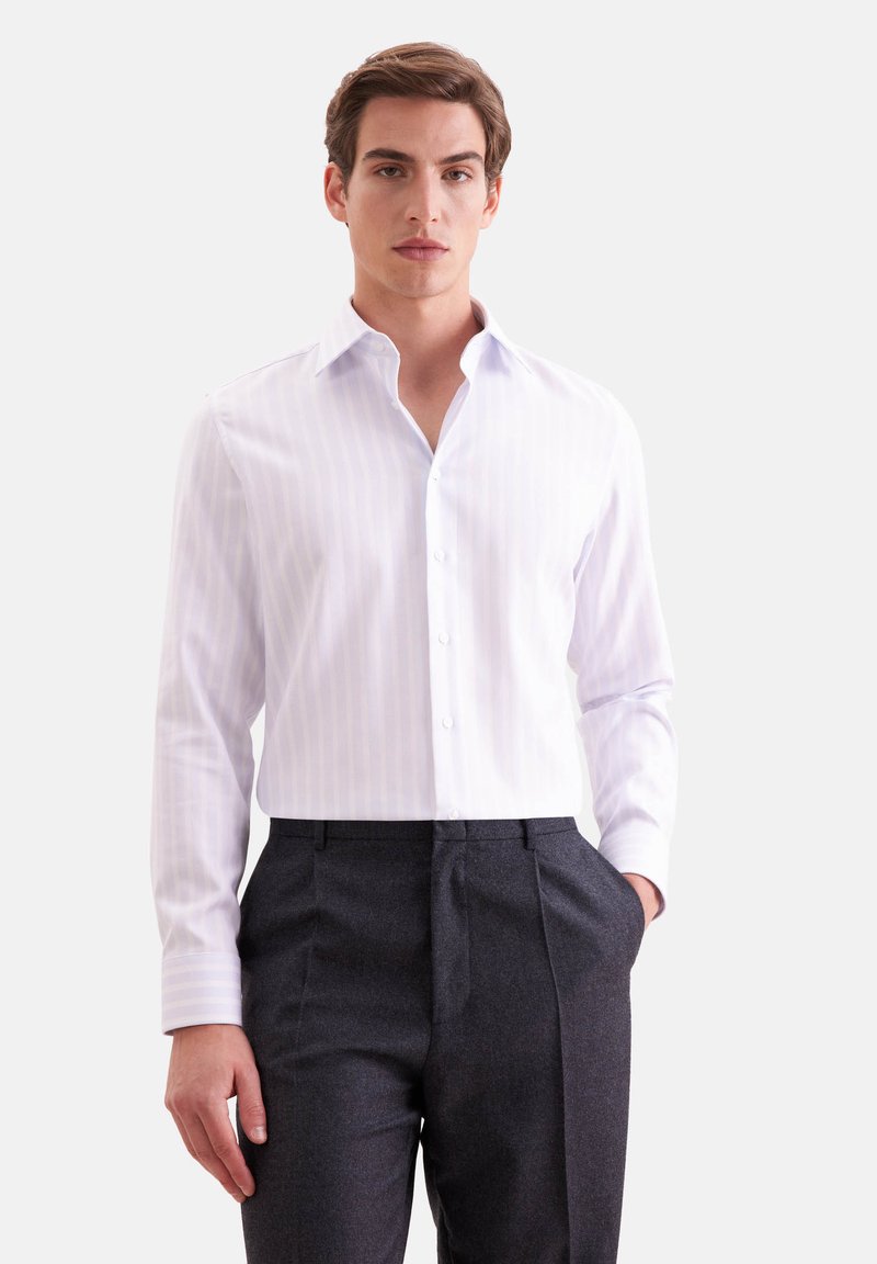Jeune homme portant une chemise blanche à manches longues boutonnée et un pantalon de costume gris foncé, debout avec une main dans la poche.